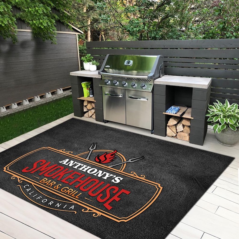 Pu&ograve; includere: Un tappeto nero da esterno con la scritta "ANTHONY'S SMOKEHOUSE BAR & GRILL CALIFORNIA" in rosso e arancione. Il tappeto &egrave; davanti a un barbecue in acciaio inossidabile e una cucina esterna in mattoni grigi. Ambiente giardino.