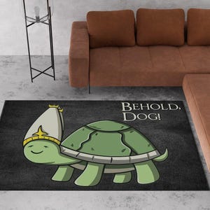 May include: A black rectangular rug featuring a cartoon turtle wearing a hat and a crown. The turtle is green with a light green shell and a white hat with a gold crown. The text "BEHOLD, DOG!" is printed in white.