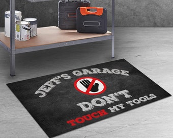 May include: A rectangular black floor mat featuring the white text JEFF'S GARAGE DON'T TOUCH MY TOOLS and a red circular prohibition symbol containing a black hand icon. The mat is positioned on a gray floor in front of a metal workbench.
