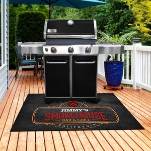 Pu&ograve; includere: Un barbecue a gas nero e argento con tre manopole di controllo e un termometro. Un tappeto nero davanti al barbecue presenta il testo "Jimmy's Smokehouse Bar & Grill California" in rosso e arancione.