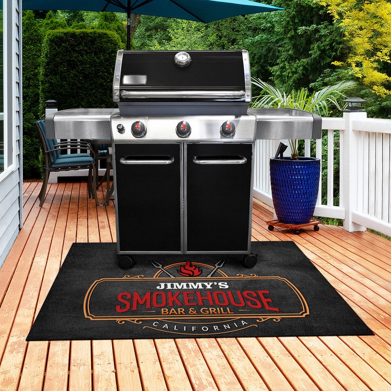Pu&ograve; includere: Un barbecue a gas nero e argento con tre manopole di controllo e un termometro. Un tappeto nero davanti al barbecue presenta il testo "Jimmy's Smokehouse Bar & Grill California" in rosso e arancione.