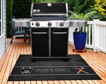 May include: A black and stainless steel gas grill with a lid-mounted thermometer and two cabinet doors. Beneath the grill sits a rectangular black floor mat featuring a skull emblem, the text JAKE'S GRILL, and a graphic of a fork and spatula.