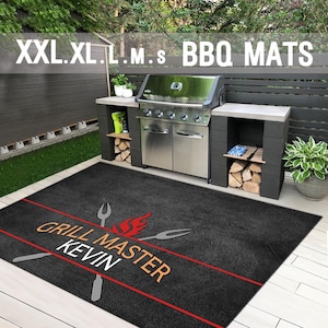 Op de afbeelding: Een rechthoekige zwarte BBQ-mat met een rode horizontale lijn, een afbeelding van twee gekruiste grillvorken, een rood vlamicoon en de tekst "GRILL MASTER KEVIN" in oranje en wit, geplaatst op een lichtgekleurd houten terras.