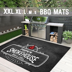 Puede incluir: Una alfombra de barbacoa negra con el texto blanco "Pope's Smokehouse Bar & Grill California". La alfombra está frente a una parrilla de acero inoxidable y una cocina exterior gris. El texto "XXL.XL.L.M.S BBQ MATS" está en la parte superior.