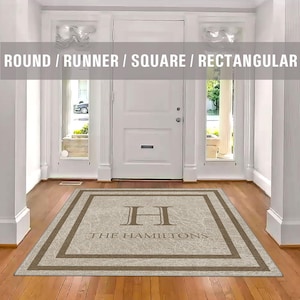 May include: A beige doormat with a brown border and the text "The Hamiltons" and a large "H" in the center. The doormat is on a wooden floor in a hallway with a white door and two windows.
