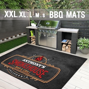 Pu&ograve; includere: Un tappeto nero per barbecue con la scritta "Anthony's Smokehouse Bar & Grill California" in rosso e arancione. Il tappeto &egrave; davanti a una griglia in acciaio inossidabile e a una cucina esterna grigio scuro. Il testo "XXL.XL.L.M.S BBQ MATS" &egrave; in alto.