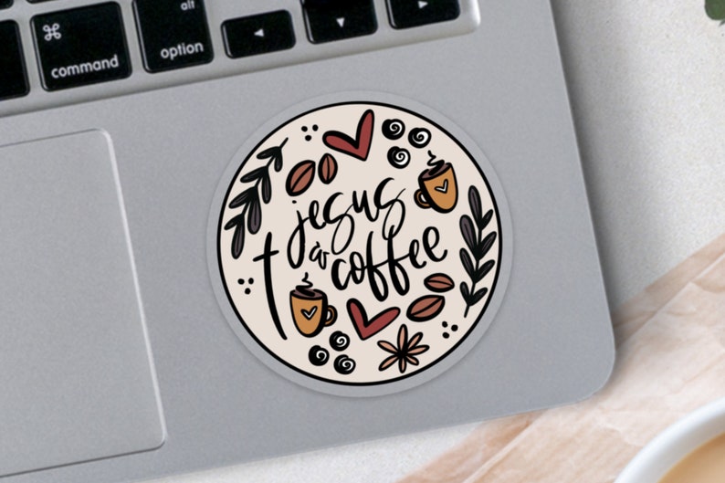 May include: A round sticker with the text "Jesus & Coffee" in a handwritten style. The design includes coffee beans, coffee cups, hearts, and floral elements in brown, black, and cream colors. The sticker is on a laptop.