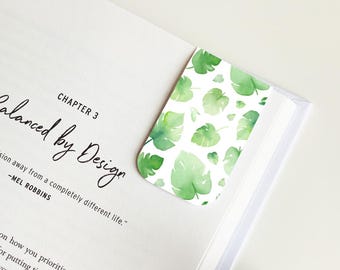 May include: A white bookmark with a green watercolor leaf pattern. The bookmark is placed on a book with the text "Chapter 3 Balanced by Design" visible. The text below reads "A decision away from a completely different life." -Mel Robbins.