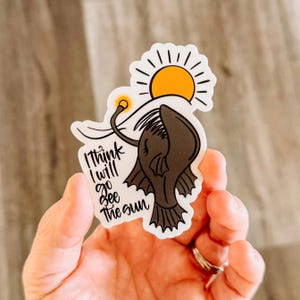 May include: A white sticker featuring a black anglerfish illustration with the text "I think I will go see the sun". The anglerfish has a glowing lure and is facing a yellow sun with rays.
