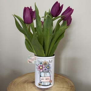 May include: A white ceramic vase with a floral design and the text "MRS SMITH" holds several purple tulips with green leaves. The vase is decorated with colorful flowers and a mason jar illustration. The vase sits on a wooden surface.
