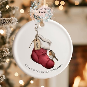 May include: A white ceramic Christmas bauble featuring a red stocking design and a small bird. The stocking has a tag that says "MUM". The text "STILL PART OF EVERY CHRISTMAS" is printed on the bottom.