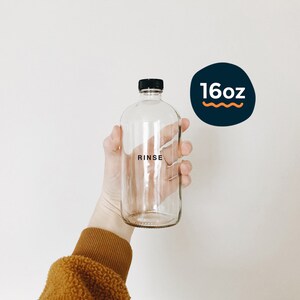 Könnte beinhalten: Eine klare Glasflasche mit schwarzem Deckel und dem Wort "RINSE" auf der Vorderseite. Die Flasche ist mit "16oz" auf einer marineblauen und orangefarbenen Grafik beschriftet. Die Flasche wird von einer Person gehalten.