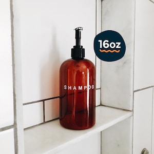 Könnte beinhalten: Eine braune Glasflasche mit einem schwarzen Pumpspender, die mit "SHAMPOO" beschriftet ist, auf einem weißen Fliesenregal. Die Flasche hat ein Fassungsvermögen von 16 Unzen.