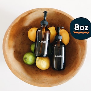 Könnte beinhalten: Zwei bernsteinfarbene Glasflaschen mit schwarzen Pumpen und weißen Etiketten. Die Etiketten lauten "HANDS" und "DISHES". Die Flaschen befinden sich in einer Holzschale mit Zitronen und Limetten.