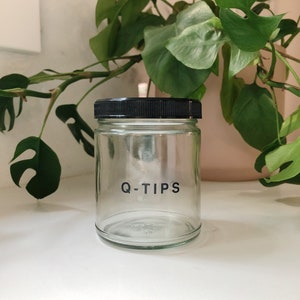 K&ouml;nnte beinhalten: Ein klares Glas mit schwarzem Deckel, beschriftet mit "Q-TIPS" in schwarzen Gro&szlig;buchstaben. Das Glas steht auf einer wei&szlig;en Oberfl&auml;che, im Hintergrund gr&uuml;ne Bl&auml;tterpflanzen.