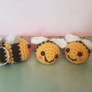 May include: Three crocheted bee keychains with black eyes and smiling mouths. Two bees are yellow and black with white wings, and one bee is yellow and black with white wings and a silver key ring.
