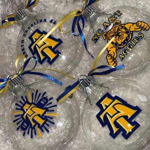 May include: Four clear glass Christmas ornaments with different designs. One ornament features the text "NC A&T Aggies" with a bulldog mascot. Another ornament features the text "North Carolina A&T" with a blue and gold letter "A". The remaining two ornaments feature a blue and gold letter "A" with a starburst design.