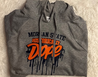 May include: Gray hoodie with the text "MORGAN STATE HISTORICALLY Dope" in navy blue and orange. The word "Dope" has a dripping effect. The hoodie has a drawstring and a hood.