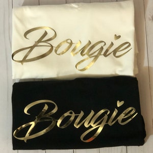 Puede incluir: Dos camisetas, una blanca y otra negra, con la palabra "Bougie" escrita en letras doradas en la parte delantera. Ambas camisetas tienen un pequeño corazón dorado al lado de la palabra.