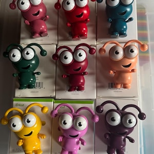 May include: Nine colorful, small, plastic figurines of a creature with two large eyes, a wide smile, and two antennae. The figurines are in various colors, including red, pink, purple, yellow, green, blue, and brown.