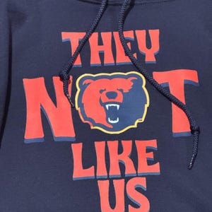 Puede incluir: Sudadera con capucha azul marino con la frase "THEY NOT LIKE US" en letras rojas. Un gráfico de oso en rojo y azul marino está centrado en la sudadera. La sudadera tiene un cordón y una capucha.