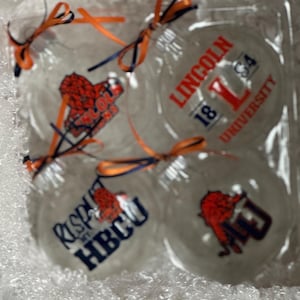 Könnte beinhalten: Vier transparente Glaskugeln mit orangefarbenen und blauen Schleifen. Die Kugeln sind mit Logos verschiedener Universitäten verziert: Lincoln University, HBCU und zwei weitere mit Logos, die nicht klar erkennbar sind.