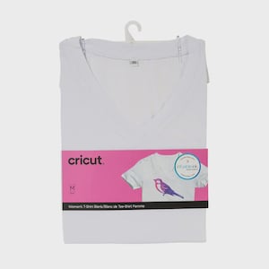 Puede incluir: Una camiseta blanca con cuello en V doblada, talla M, con una etiqueta rosa con el logotipo de Cricut y un gráfico de un pájaro en una camiseta. La etiqueta también incluye el texto "Women's T-Shirt Blank".