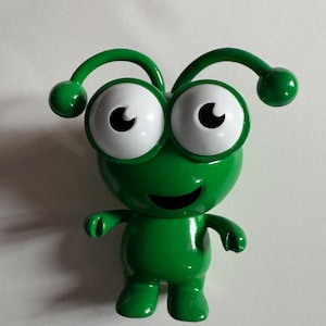 以下が含まれることがあります： 白い大きな目で笑顔の緑色のエイリアンの玩具。エイリアンは2本のアンテナがあり、白い表面の上に立っています。