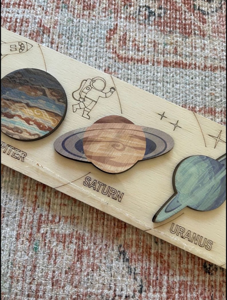 Puede incluir: Un rompecabezas de madera con los planetas J&uacute;piter, Saturno y Urano. Los planetas se representan en un dise&ntilde;o estilizado y colorido con anillos alrededor de Saturno y Urano. El rompecabezas tambi&eacute;n incluye una peque&ntilde;a figura de astronauta.