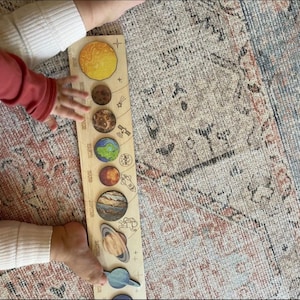 Puede incluir: Un rompecabezas de madera del sistema solar con planetas coloridos y una mano de ni&ntilde;o colocando la &uacute;ltima pieza. El rompecabezas incluye los nombres de cada planeta.