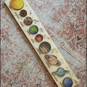 Puede incluir: Una tabla de madera con un sistema solar pintado, que muestra el sol y los planetas en orden desde el sol: Mercurio, Venus, Tierra, Marte, J&uacute;piter, Saturno, Urano y Neptuno. Cada planeta es una pieza de madera separada con una imagen pintada del planeta.
