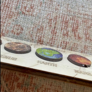 Puede incluir: Primer plano de un modelo de sistema solar de madera con planetas pintados. Los planetas est&aacute;n etiquetados como "Venus", "Tierra" y "Marte".