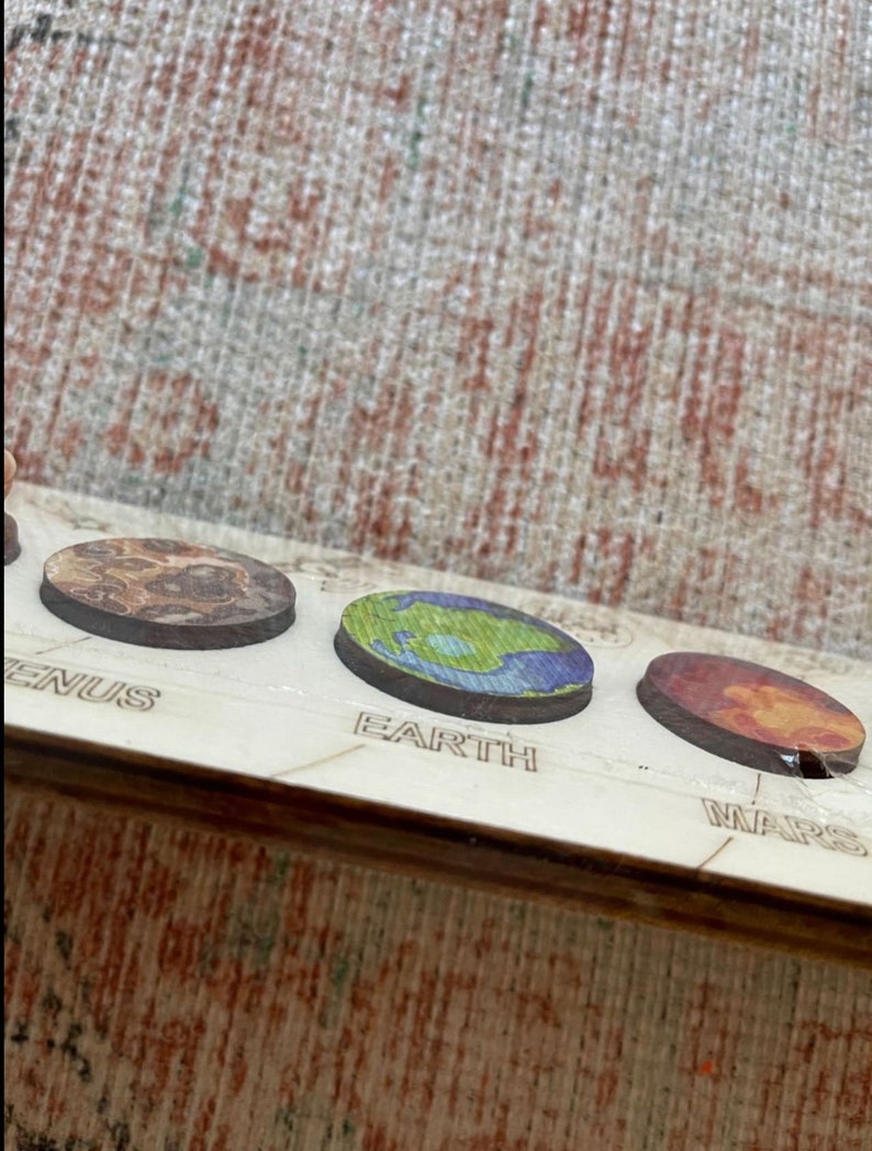 Puede incluir: Primer plano de un modelo de sistema solar de madera con planetas pintados. Los planetas est&aacute;n etiquetados como "Venus", "Tierra" y "Marte".