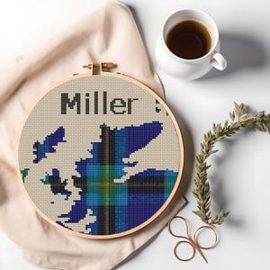 Pode incluir: Um bastidor de bordado em ponto cruz com o nome "Miller" acima de um desenho xadrez azul e verde. O bastidor está sobre um tecido bege, com uma chávena de chá, um raminho de verdura e tesouras por perto.
