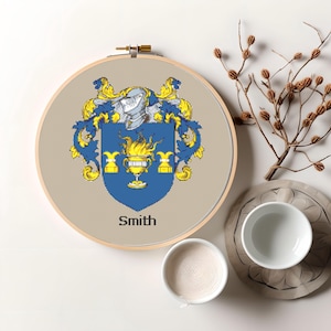 May include: A round embroidery hoop with a beige background featuring a blue and gold coat of arms with the name "Smith" printed below. The coat of arms has a blue shield with a gold flame and a gold chalice. The shield is surrounded by gold floral designs.