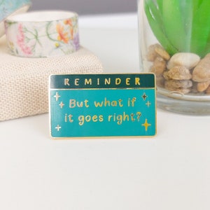 Pode incluir: Um alfinete retangular de esmalte com fundo turquesa e borda dourada. O alfinete diz "REMINDER" no topo e "But what if it goes right?" em letras douradas. H&aacute; estrelas decorativas no alfinete.