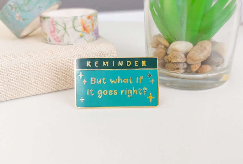 Pode incluir: Um alfinete retangular de esmalte com fundo turquesa e borda dourada. O alfinete diz "REMINDER" no topo e "But what if it goes right?" em letras douradas. H&aacute; estrelas decorativas no alfinete.