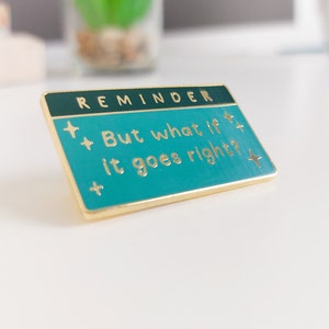 Pode incluir: Um alfinete retangular de esmalte com fundo turquesa e borda dourada. O alfinete diz "REMINDER" no topo e "But what if it goes right?" em letras turquesas. O alfinete tem detalhes de estrelas douradas.