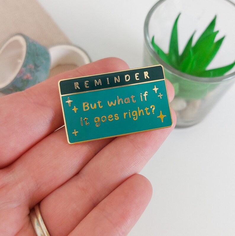 Pode incluir: Um alfinete de esmalte retangular com fundo turquesa e borda dourada. O alfinete diz "REMINDER" na parte superior e "But what if it goes right?" em letras douradas. O alfinete tem detalhes de estrelas douradas e &eacute; segurado em uma m&atilde;o.