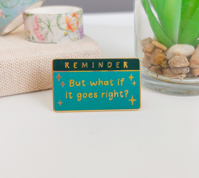 Pode incluir: Um alfinete retangular esmaltado com fundo turquesa e borda dourada. O alfinete diz "REMINDER" no topo e "But what if it goes right?" em letras douradas. Explos&otilde;es de estrelas douradas nas laterais. Fita adesiva floral no fundo.