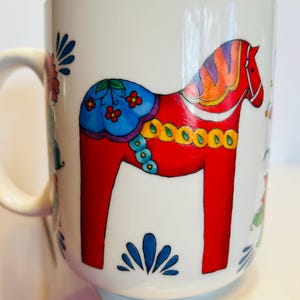 May include: White ceramic mug featuring a vibrant red Dala horse design with blue, yellow, and orange accents. The mug also has floral and leaf patterns. A classic and colorful piece for coffee or tea.
