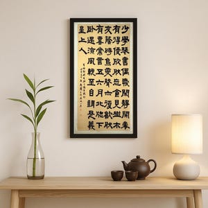 Könnte beinhalten: Ein gerahmtes Kunstwerk mit chinesischer Kalligraphie in schwarzer Tinte auf goldenem Hintergrund. Der schwarze Rahmen umgibt den vertikalen Text. Das Kunstwerk wird auf einem Holztisch mit einer Teekanne, Tassen und einer Lampe ausgestellt.