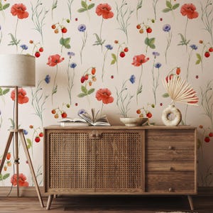 May include: A room featuring a floral wallpaper design with red poppies, strawberries, and blue cornflowers. A wooden cabinet with a woven front and a lamp with a beige shade are in the foreground. A vase with a dried palm leaf and a bowl are on the cabinet.