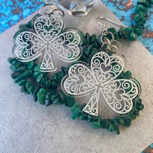 May include: A pair of clear acrylic earrings with a white, intricate shamrock design. The earrings are displayed on a green stone necklace.