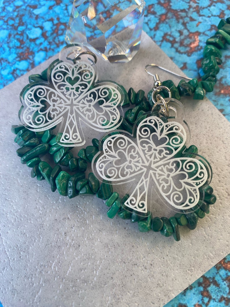 May include: A pair of clear acrylic earrings with a white, intricate shamrock design. The earrings are displayed on a green stone necklace.