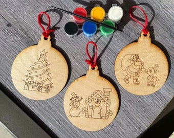 May include: Three unfinished wooden Christmas ornaments with red string hangers. The ornaments are round and feature different designs: a Christmas tree, Santa Claus with a reindeer, and a snowman with a gingerbread house. The ornaments are ready to be painted with the included set of six paint pots and a paintbrush.