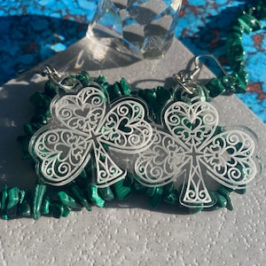 May include: A pair of clear acrylic earrings shaped like shamrocks with a white filigree design. The earrings are on a green malachite background.