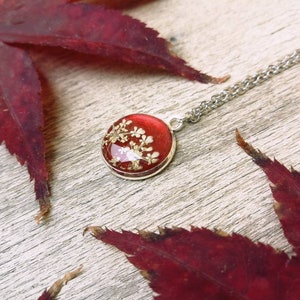Puede incluir: Un collar de cadena de plata con un colgante rojo y dorado. El colgante es una pieza redonda de resina transparente con flores doradas prensadas en su interior.