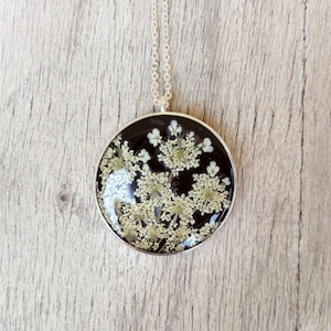 Black resin pendant with white wildflowers inserted. Diameter 3.3 cm, thickness about 7 mm. Thin chain in stainless steel.