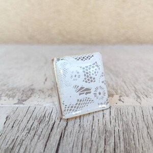 Può includere: Un anello quadrato bianco con un delicato motivo a pizzo. L'anello è realizzato in resina e ha una finitura lucida.